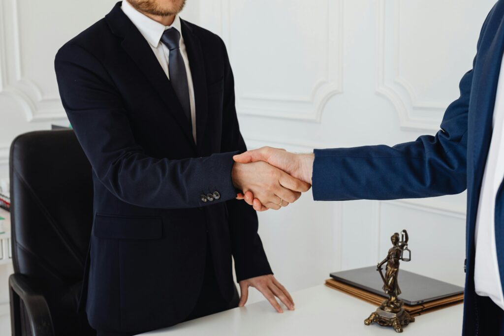 Two men shaking hands in a professional setting, symbolizing trustworthy immigration advice. The image represents Morgan Smith Immigration's commitment to ethical services amid new UK government reforms cracking down on rogue advisers. Background documents and a laptop suggest visa application reviews in line with Home Office regulations under the Border Security Bill