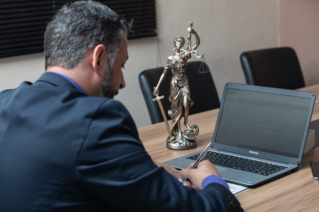Lawyer working on a laptop with Lady Justice statue beside him, symbolising legal processes and debates on Indefinite Leave to Remain in the UK.
